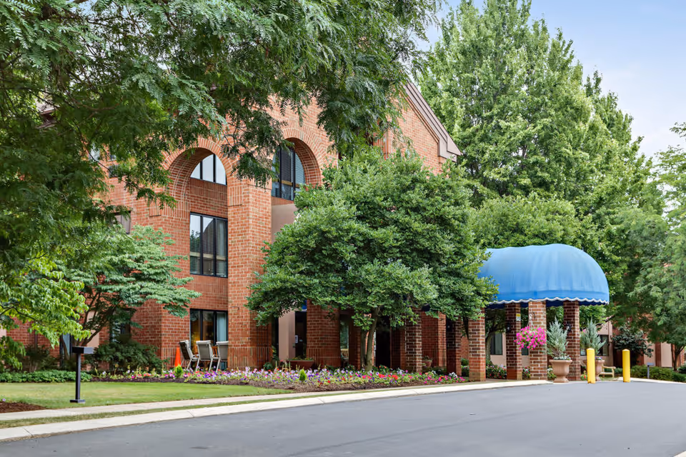 Exterior view of a brick building with arched windows partially obscured by green trees and shrubs. There is a blue canopy over the entrance, surrounded by flower beds and a paved driveway in front.