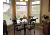 A small dining area with a round glass table and four wrought iron chairs with cushions, situated in a bay window nook with white blinds. Outside the windows, greenery and a brick building are visible. There is a wooden cabinet with a flower arrangement on the right side.