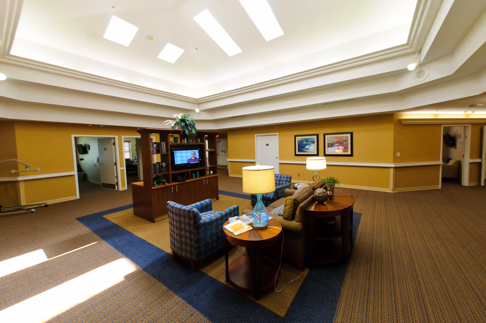 A spacious and well-lit common area with a high ceiling and skylights. The room features a central wooden entertainment unit with a television and shelves holding books and decorative items. Surrounding the unit are comfortable seating options including a sofa and patterned armchairs, along with side tables holding lamps and magazines. The walls are painted mustard yellow with white trim, and there are framed paintings hanging on the wall. Several doorways lead to other rooms.