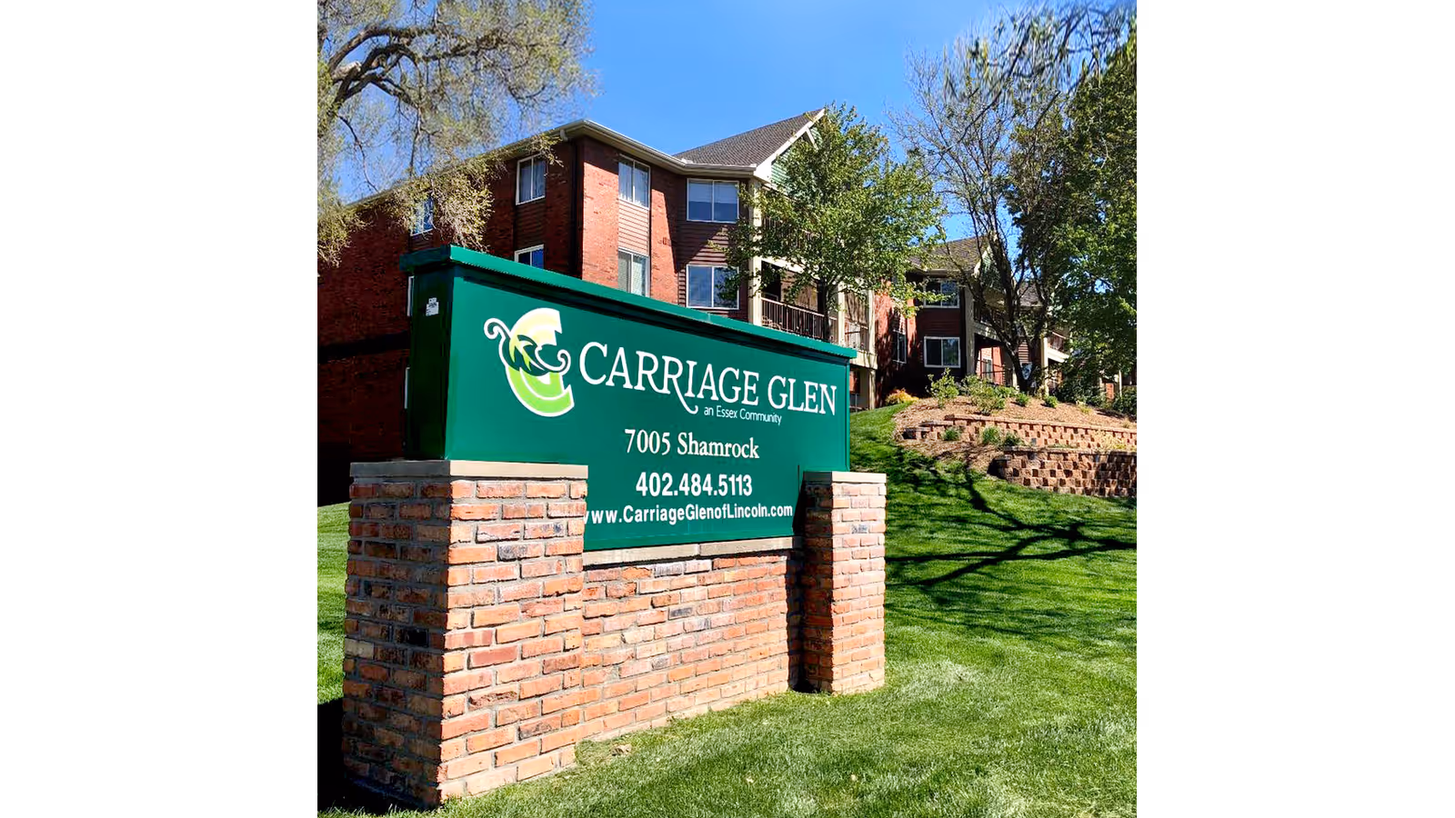 Outdoor view of a green sign for Carriage Glen senior living community, mounted on a brick base with a multi-story brick building and trees in the background under a clear blue sky.