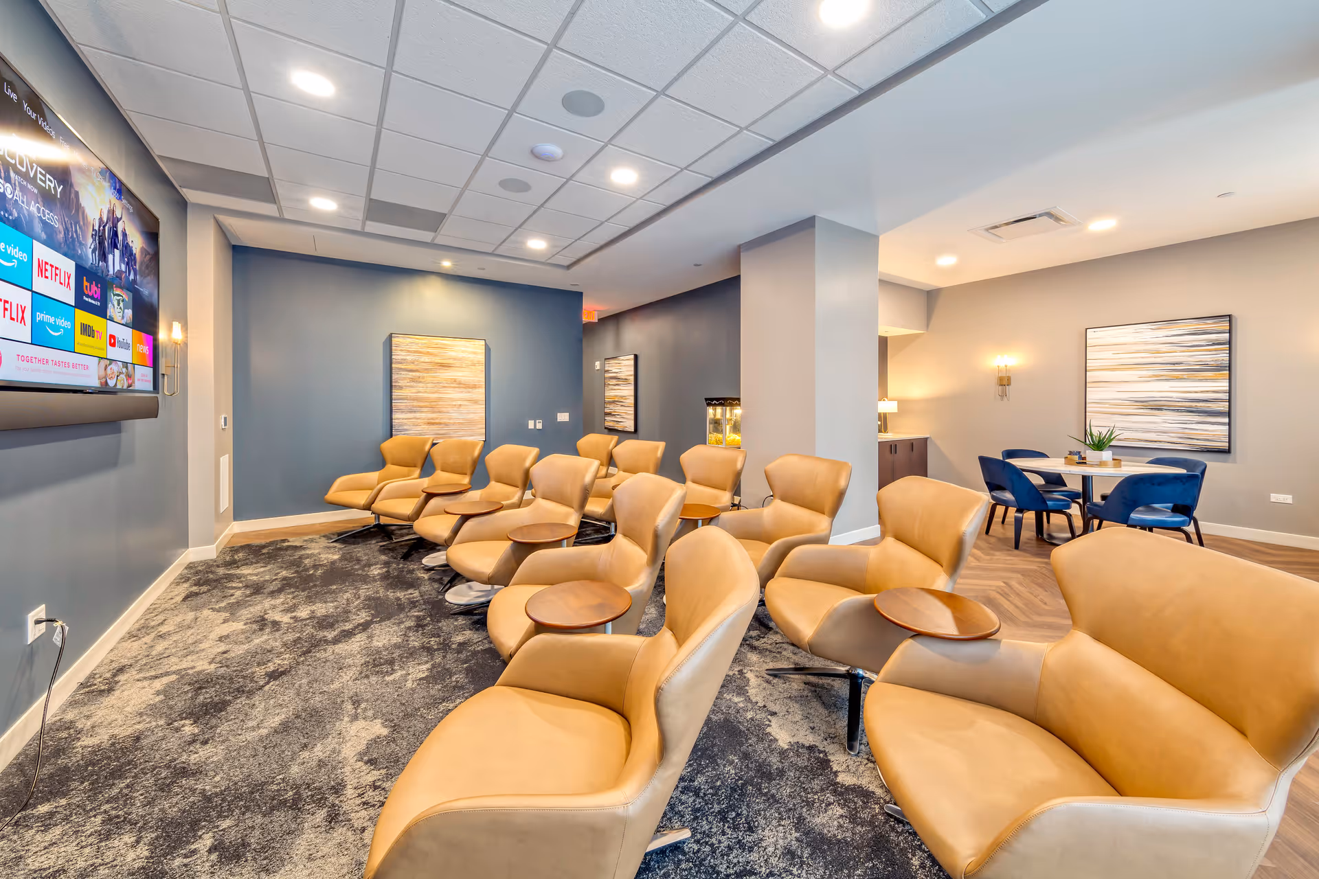 A modern lounge area with multiple tan leather chairs arranged in rows facing a large wall-mounted TV screen displaying streaming service icons. The room has blue and gray walls, carpeted flooring, and contemporary artwork on the walls. In the background, there is a small dining area with a round table and blue chairs.