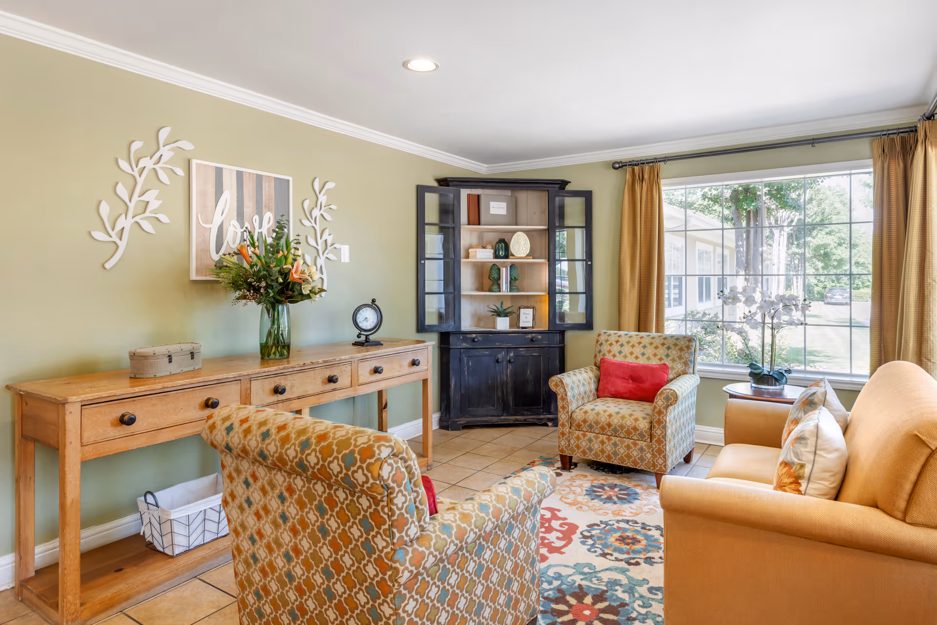 Sunlit living room with patterned armchairs, a sofa, a wooden console table with flowers, and a display cabinet by a large window.