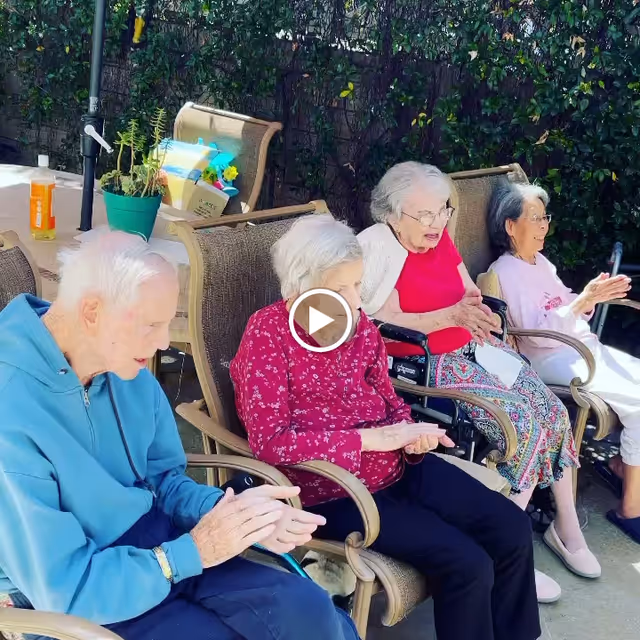Four elderly individuals sitting outdoors on patio chairs, clapping and enjoying a sunny day. They are positioned in a row with greenery in the background and a table with a potted plant and a bottle nearby.