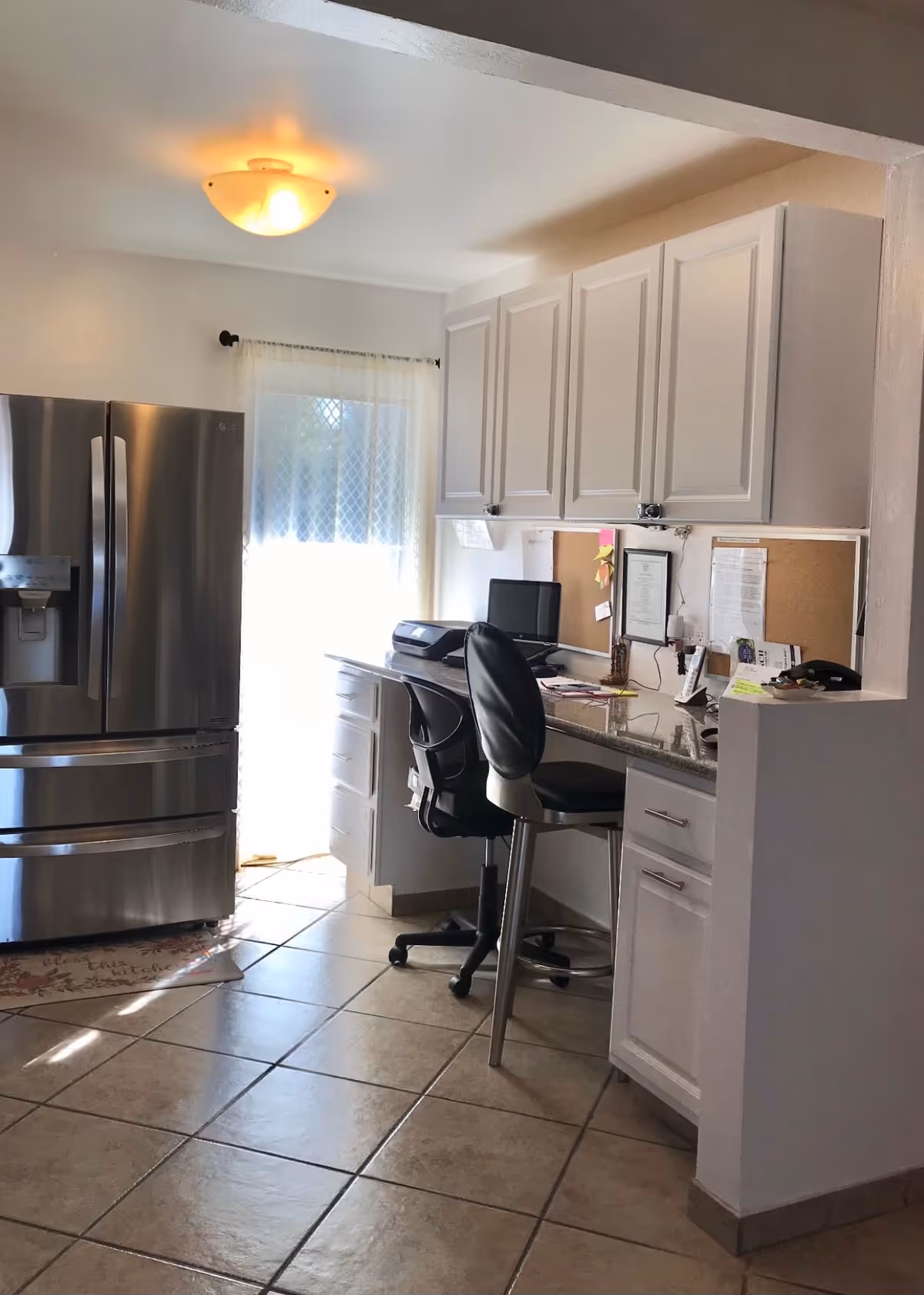 Kitchen area with a stainless steel refrigerator, white cabinets, tiled floor and a small desk nook with a chair.