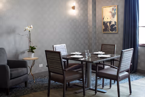 A small dining area with a square table set for four, upholstered chairs, a gray armchair, potted orchid on a side table, patterned wallpaper and framed artwork.