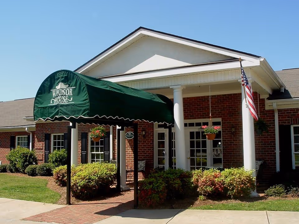 Exterior view of a brick building with a white porch supported by columns. There is a green awning with the text 'Windsor House' over the entrance. Hanging flower pots and an American flag are visible near the entrance. The surrounding area has neatly trimmed bushes and a clear blue sky.