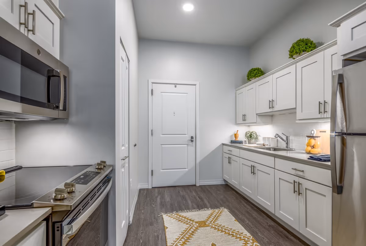 Modern kitchen with white cabinets, stainless steel appliances including a microwave, stove, and refrigerator. The kitchen has a wooden floor, a white door at the end, a small rug with a geometric pattern, and decorative green plants on top of the cabinets.