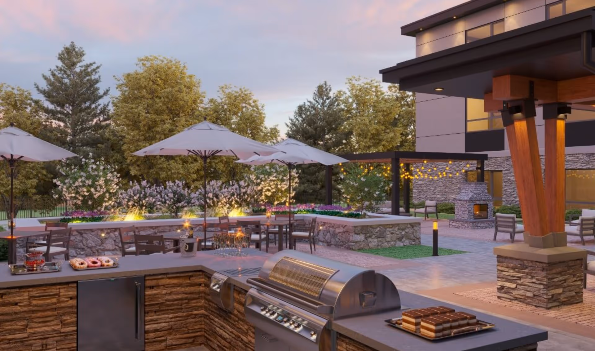 Outdoor patio area at Benchmark at Hanover featuring a built-in grill and countertop with desserts, several tables with umbrellas, stone walls, string lights, and a stone fireplace surrounded by chairs, with trees and flowering plants in the background during dusk.