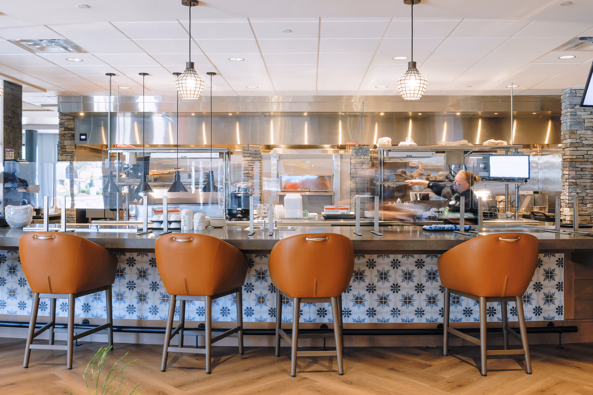 A modern dining counter with four leather bar stools facing an open commercial kitchen and pendant lights.