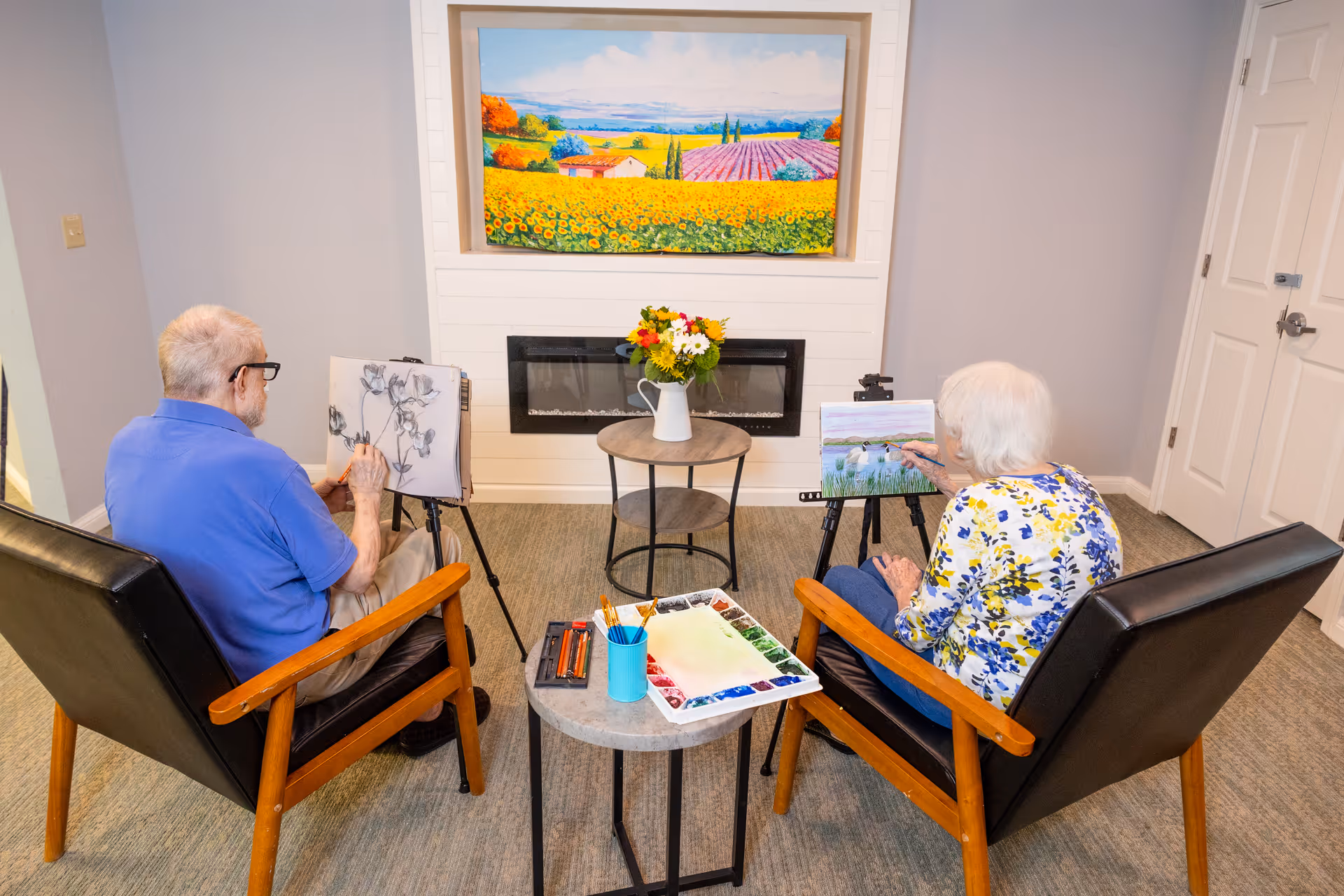 Two elderly individuals seated in armchairs facing away from the camera, engaged in painting on easels in a cozy room with a fireplace and a colorful landscape painting above it. A small table between them holds painting supplies and a vase with flowers.