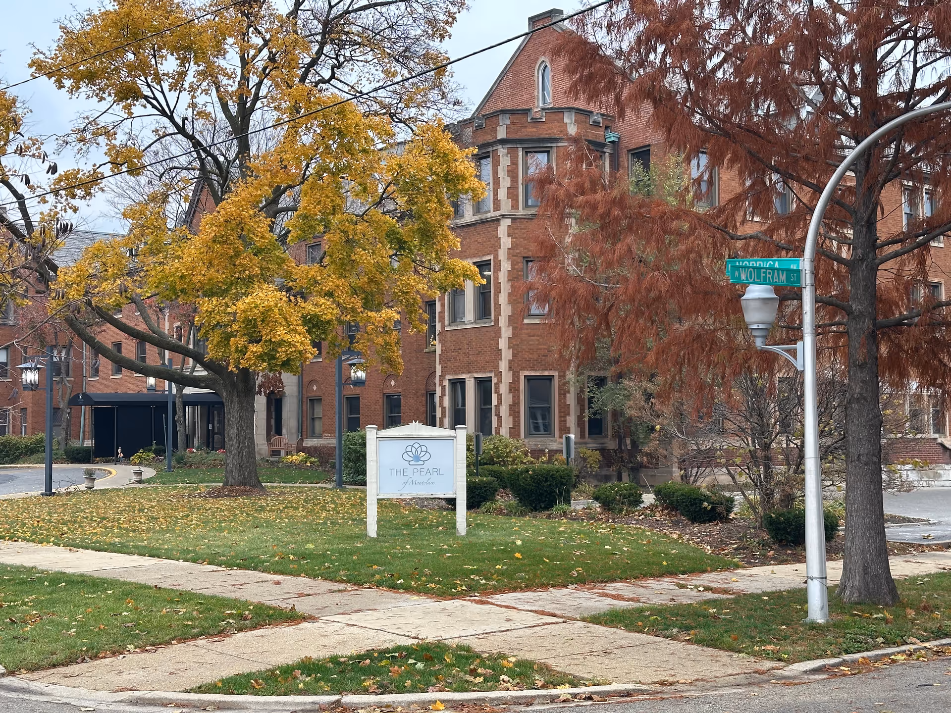 Exterior view of a brick building with autumn trees and a lawn in front. A white sign on the lawn reads 'The Pearl of Montclare'. There are street signs on a pole indicating the intersection of Nordica Ave and Wolfram St.