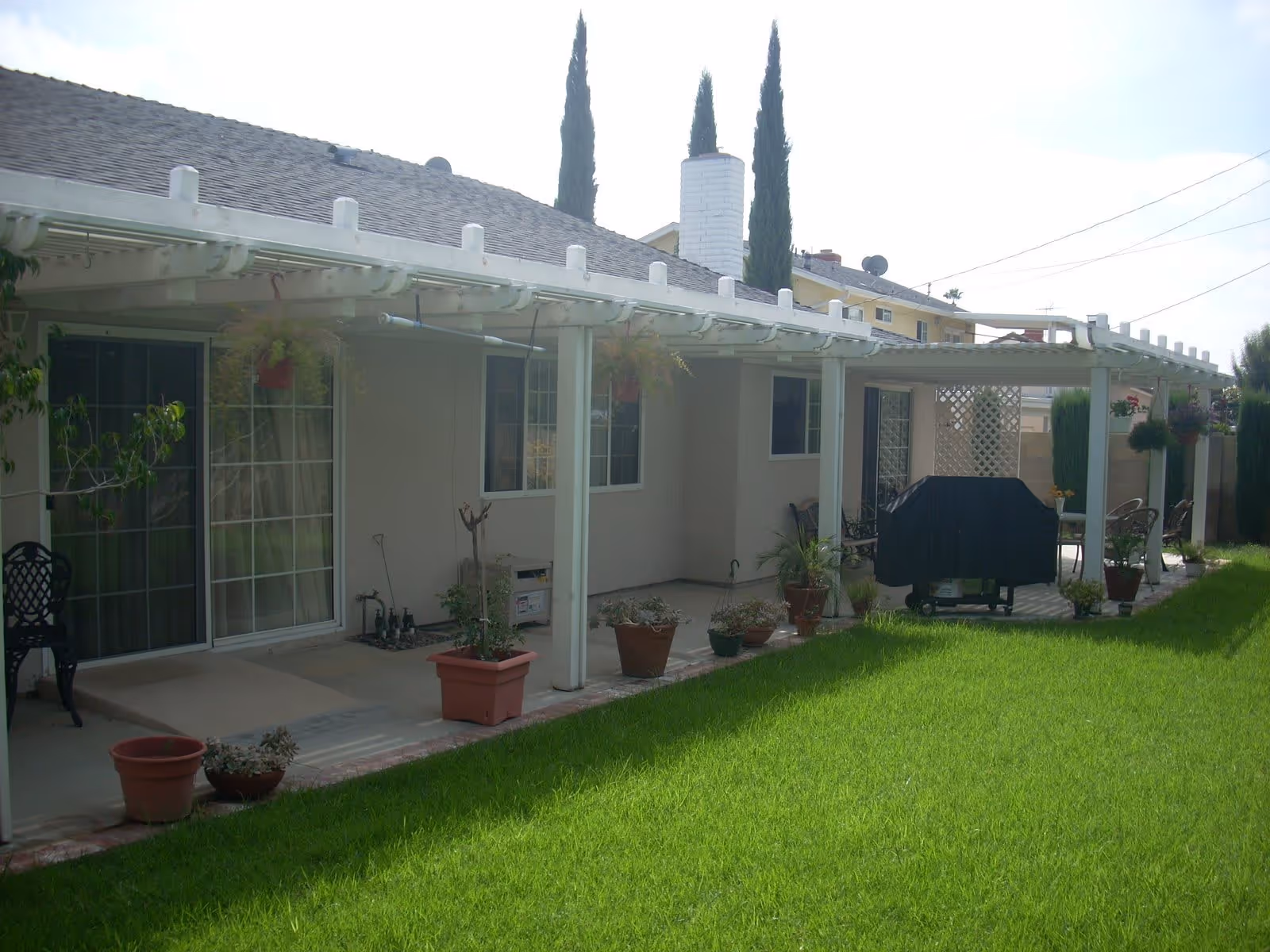 Backyard patio area of a residential building with a white pergola covering a concrete patio. Several potted plants and hanging plants are placed along the patio. There is a covered grill and outdoor seating visible under the pergola. The lawn in front is green and well-maintained.