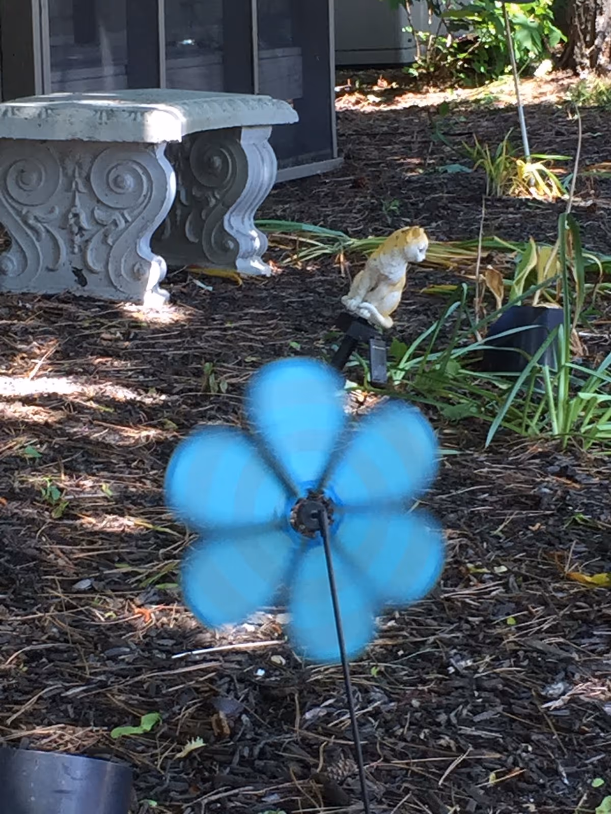 A blue flower-shaped pinwheel spins in a mulched garden bed with a decorative concrete bench and a small animal statue in the background.