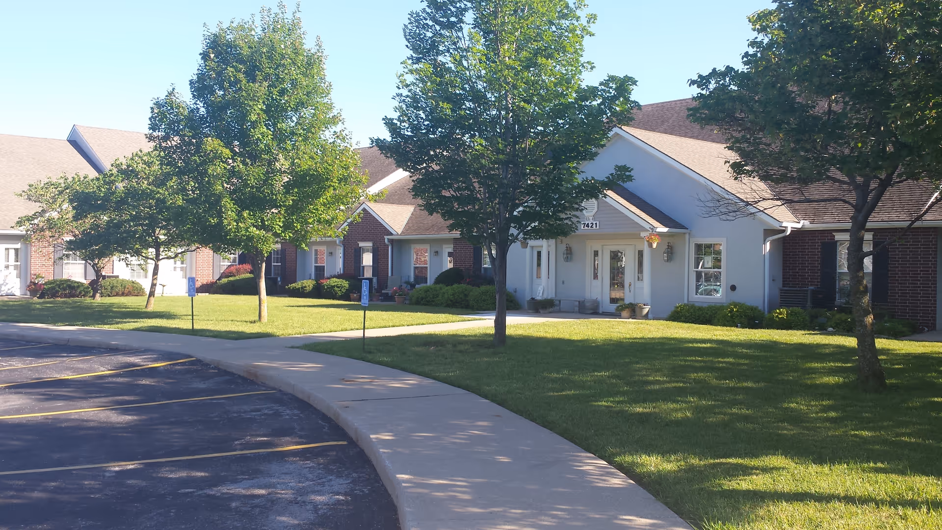 Exterior view of Elliott Place Senior Living showing a single-story building with a combination of brick and white siding, surrounded by green grass and trees, with a curved sidewalk and parking spaces in the foreground.