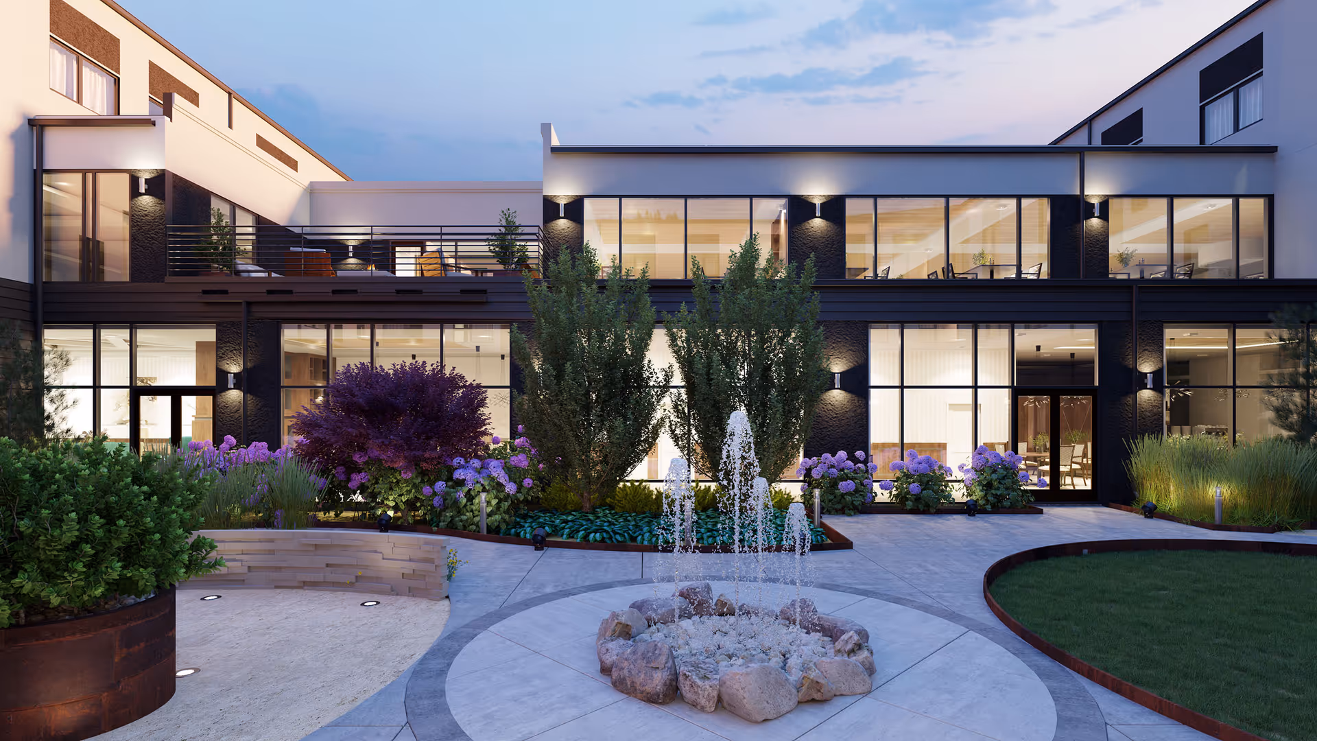 Outdoor courtyard area of a senior living facility at dusk featuring a central water fountain surrounded by rocks, landscaped greenery, purple flowers, and bushes. The building has large glass windows with interior lighting visible, and an upper balcony with seating.