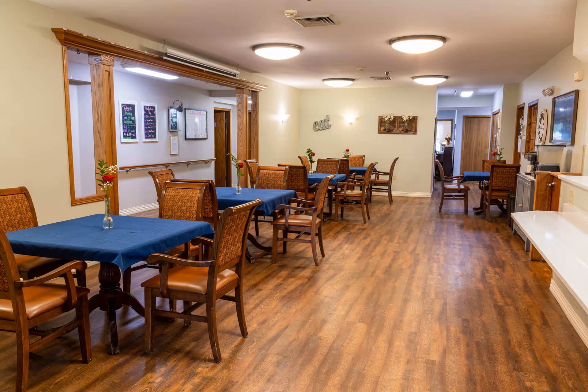 A dining room in a senior living facility with several wooden tables covered with blue tablecloths and surrounded by wooden chairs with cushioned seats. Each table has a small vase with a single red rose. The room has wood flooring, soft lighting from ceiling fixtures, and light-colored walls decorated with framed pictures and wall art. There is a counter on the right side and a hallway leading to other rooms in the background.