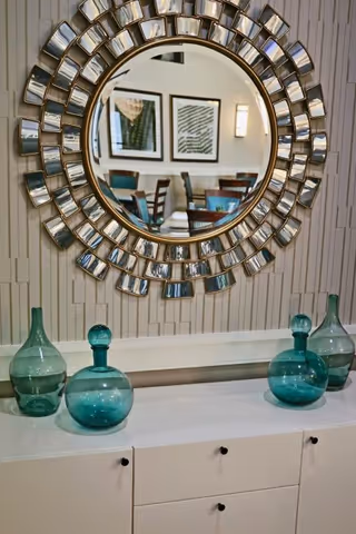 A decorative sunburst mirror hangs above a white sideboard topped with teal glass vases, reflecting a dining area with framed art and chairs.