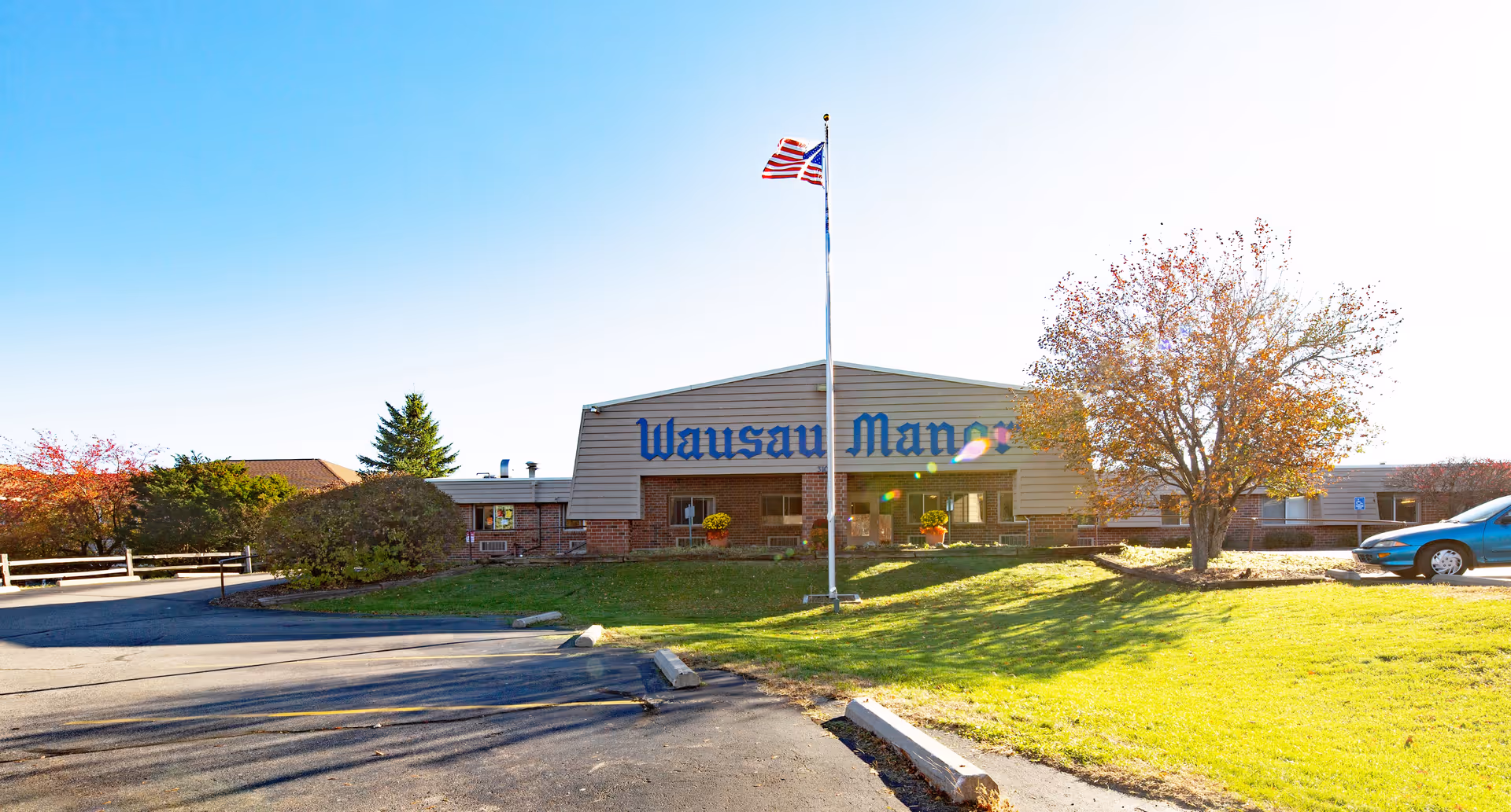Exterior view of Wausau Manor Health Services building with a flagpole flying the American flag in front, surrounded by green grass, trees with autumn leaves, and a parked car on the right side.