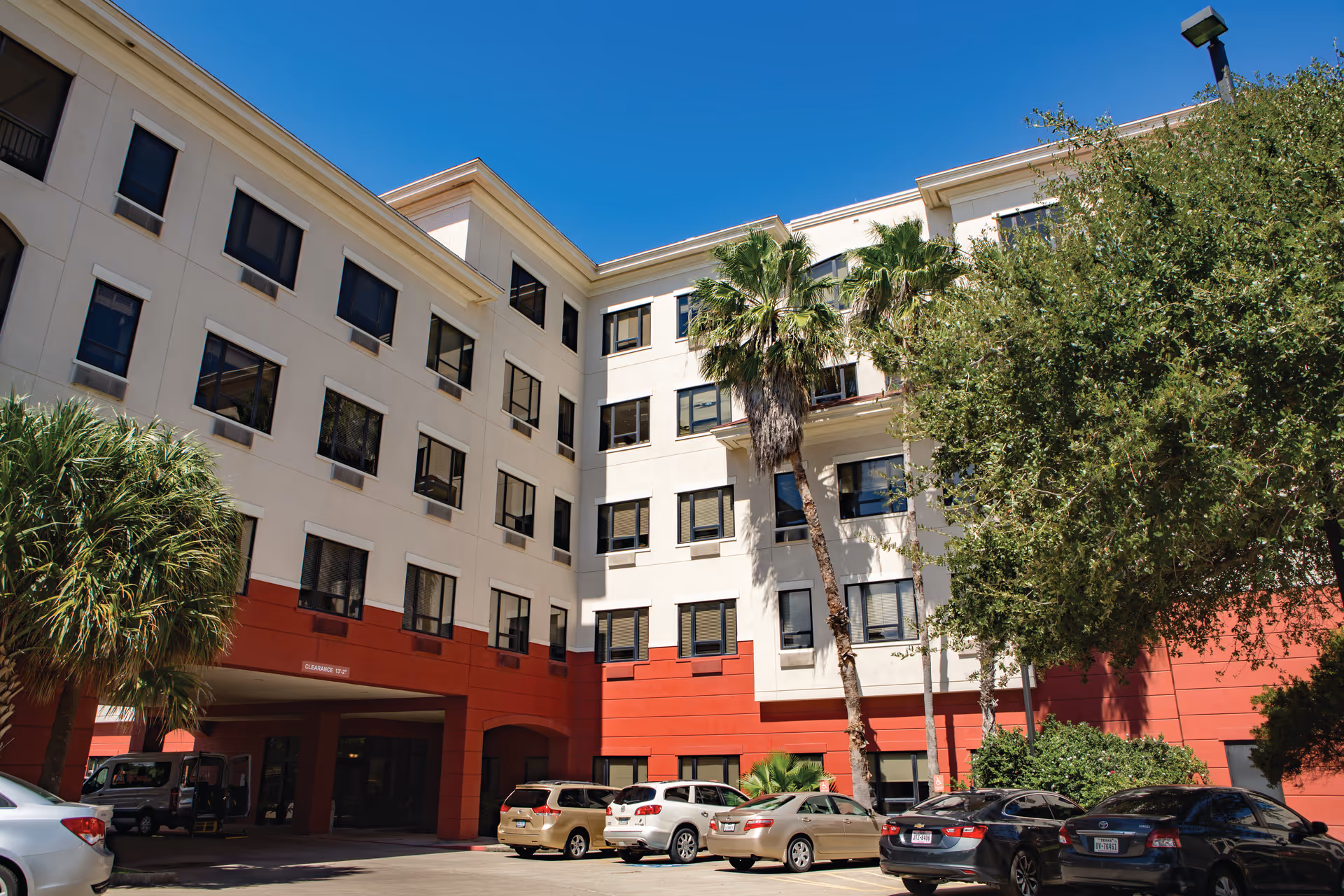 Exterior view of a multi-story senior living facility building with beige and red walls, several windows, palm trees, and parked cars in front under a clear blue sky.