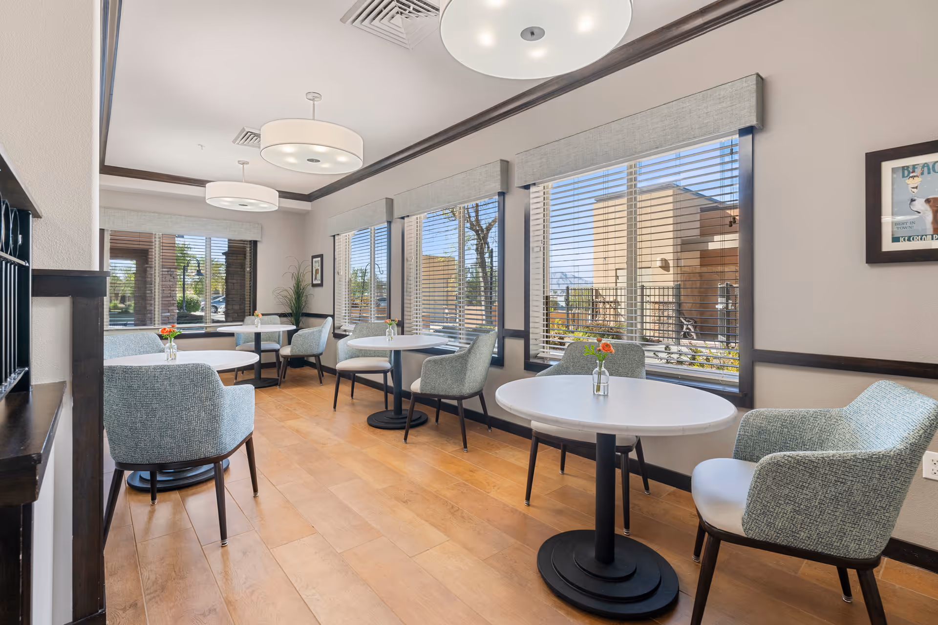 A bright and airy dining area with round white tables and comfortable upholstered chairs. The room features large windows with blinds, allowing natural light to fill the space. Small vases with flowers are placed on each table. Modern ceiling lights hang from the ceiling, and the floor is made of light wood.