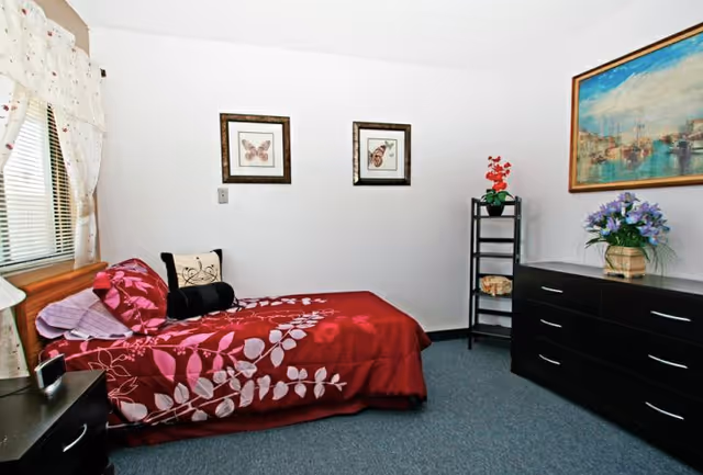 Small assisted-living bedroom with a single bed in red floral bedding, a dark dresser, framed pictures on the wall, and a window with curtains.