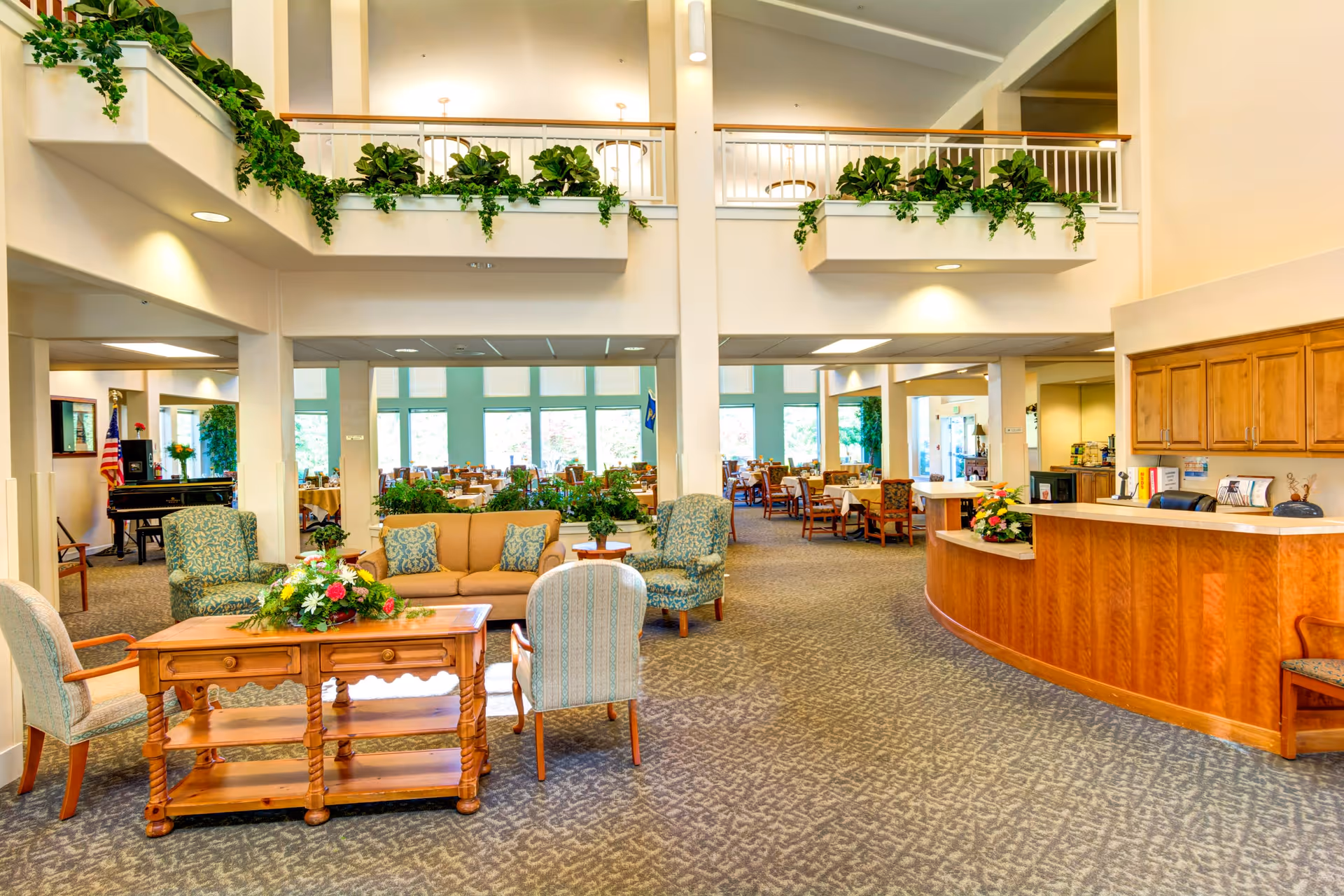 Spacious and well-lit common area in a senior living facility featuring comfortable seating with armchairs and a sofa around a wooden coffee table adorned with a flower arrangement. The area includes a reception desk on the right side and a dining area with multiple tables and chairs in the background. Green plants decorate the upper balcony railings, adding a touch of nature to the bright interior space.