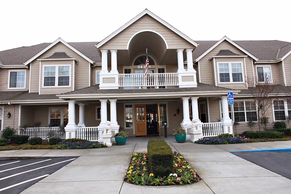 Front exterior view of a two-story senior living facility building with beige siding, white columns, and a covered entrance. There is a small garden with flowers and shrubs in front of the entrance, and an American flag hanging above the doorway. A handicap parking sign is visible near the entrance.