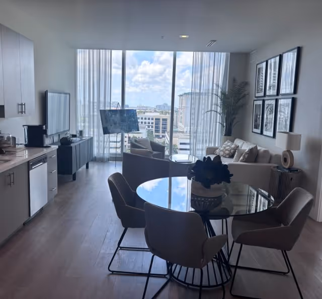 Open-plan living and dining area with a round glass table, seating, kitchenette on the left, and floor-to-ceiling windows with a city view.