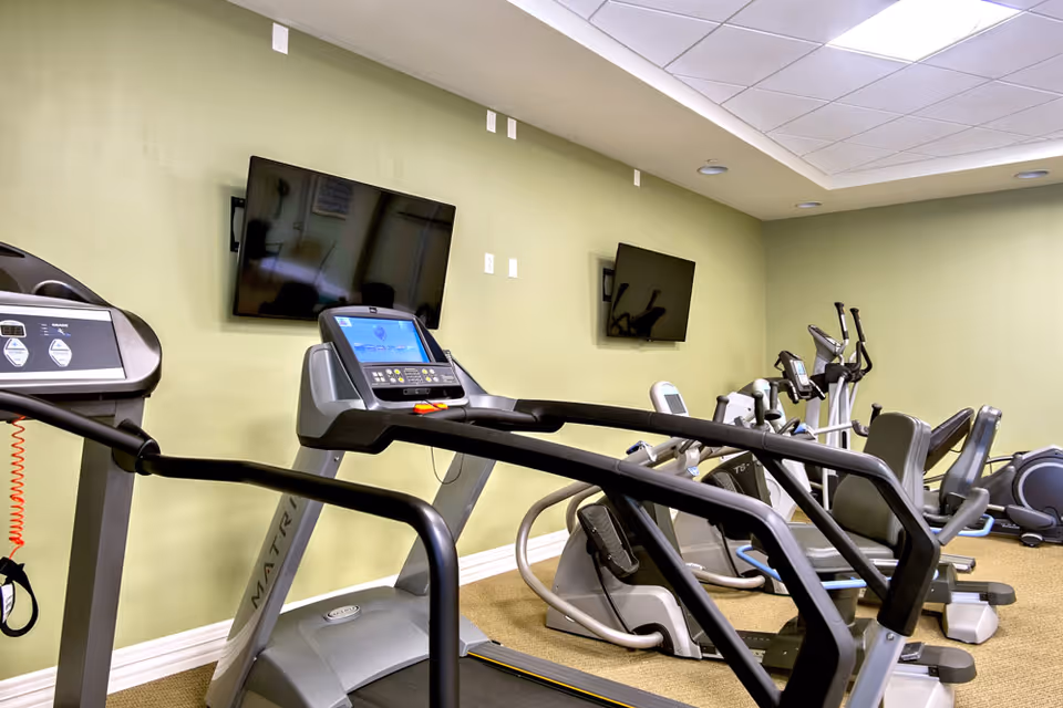 Indoor fitness room with several exercise machines including treadmills and stationary bikes, two wall-mounted flat screen TVs, and light green walls with a drop ceiling.