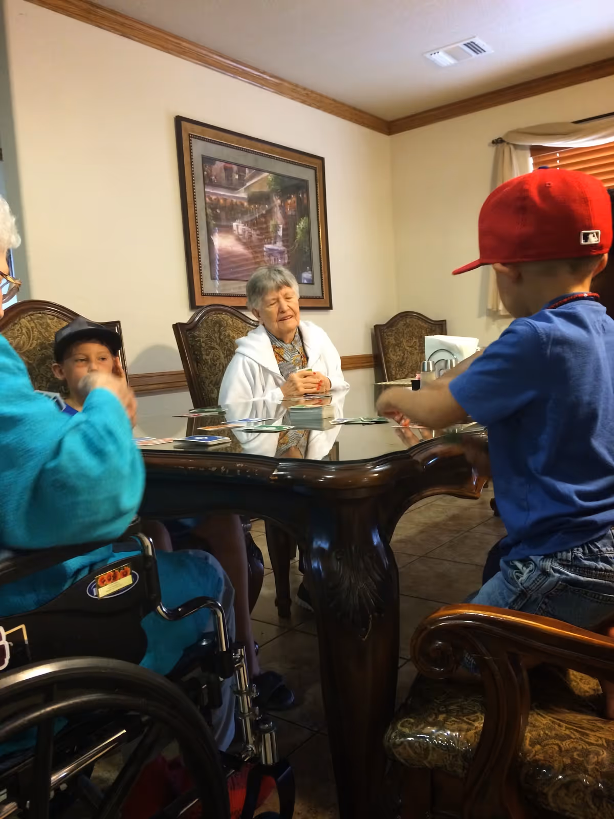 An elderly woman and two children sitting around a wooden table playing a card game in a room with beige walls and a framed picture on the wall. One child is wearing a red cap and blue shirt, and the elderly woman is wearing a white jacket. Another person in a wheelchair is partially visible.