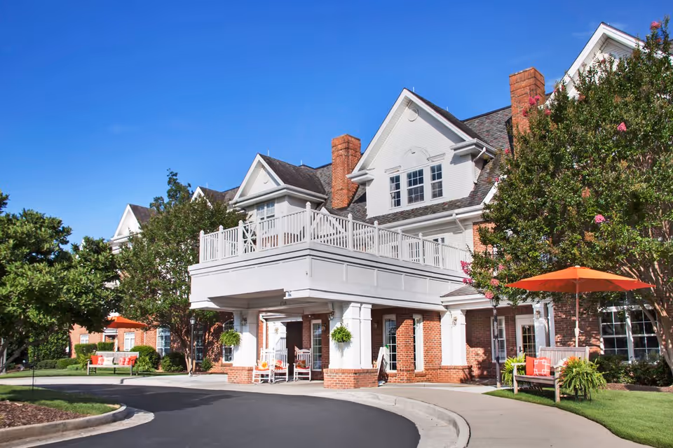 Exterior view of a senior living facility building with white siding and brick accents, featuring a covered entrance with white columns, rocking chairs, and hanging plants. There are trees and benches with orange umbrellas on a sunny day with a clear blue sky.