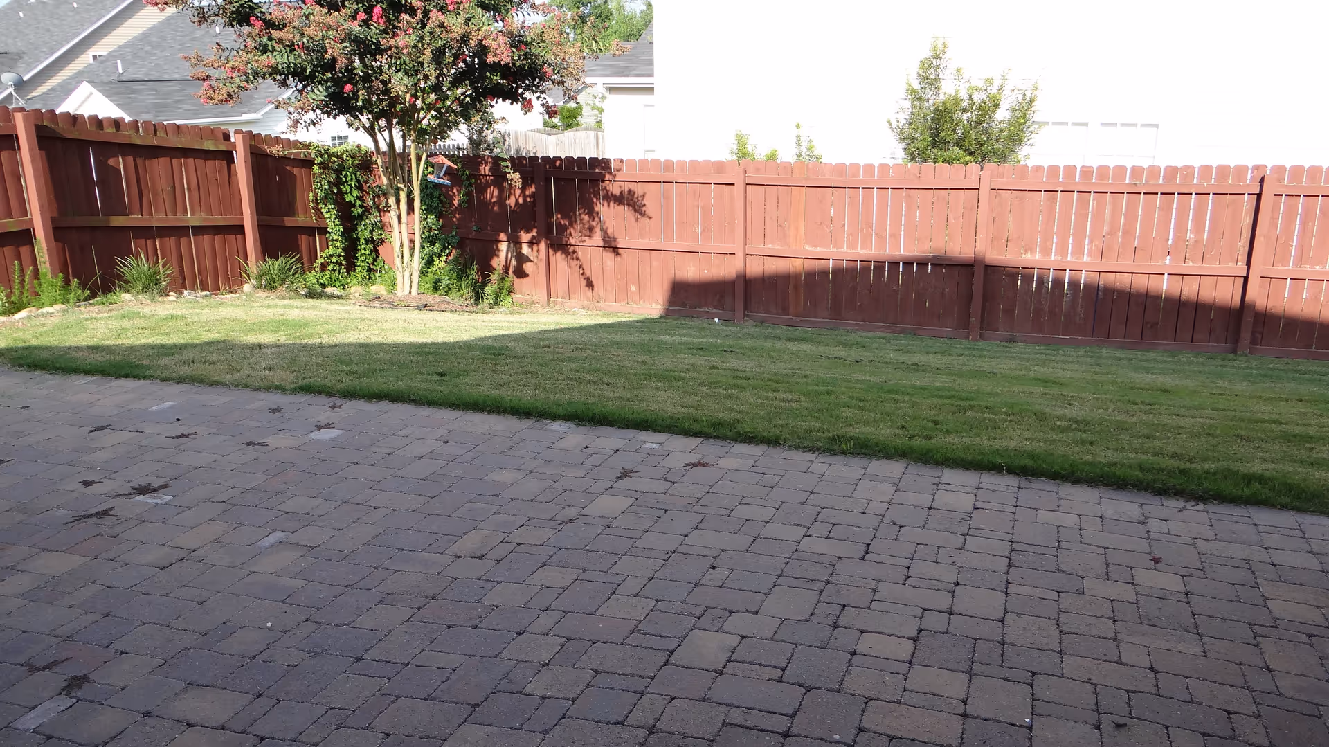 Paved patio leading to a grassy backyard bordered by a red wooden fence and a small tree.