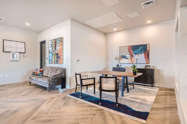 Modern communal lounge featuring a dining table and chairs on a rug, a sofa, artwork, and herringbone wood flooring.