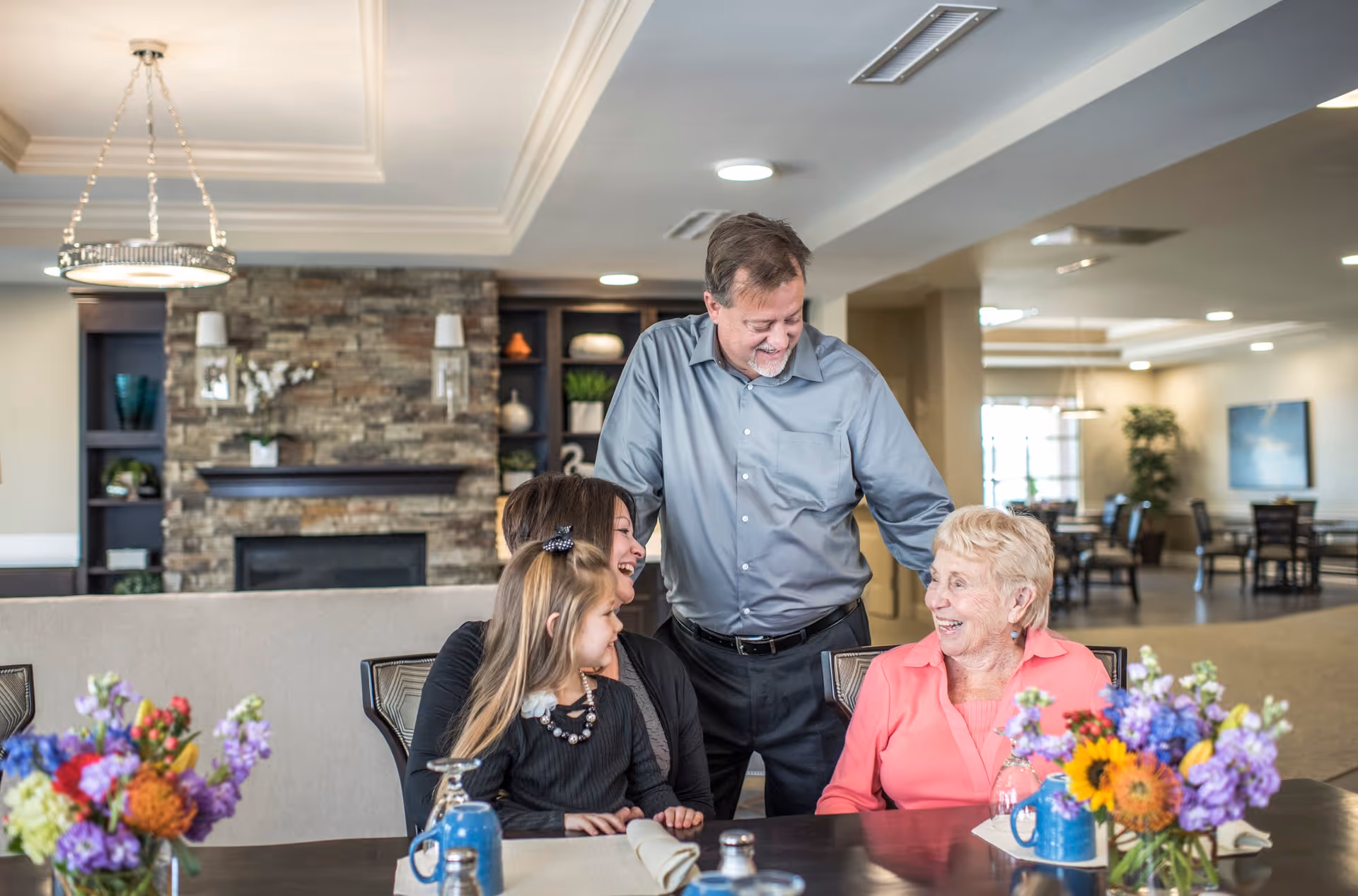An elderly woman, a middle-aged man, a middle-aged woman, and a young girl are sitting and standing around a dining table in a warmly lit room with a stone fireplace and built-in shelves in the background. The group is smiling and engaging happily with each other. The table is decorated with colorful flower arrangements and place settings.