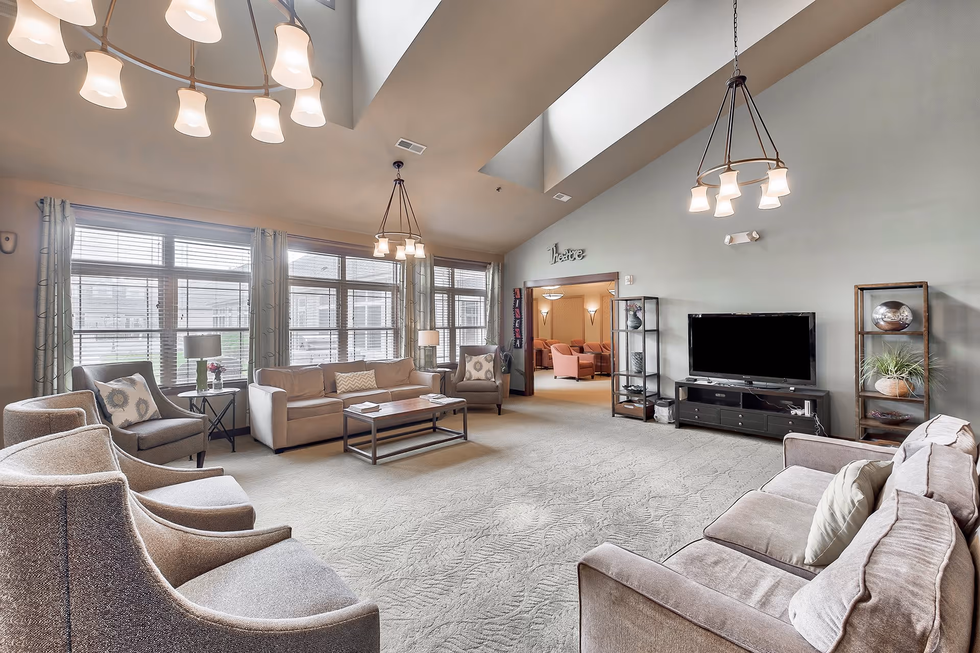 A spacious living room with high ceilings and large windows letting in natural light. The room is furnished with multiple beige and gray sofas and armchairs arranged around a central coffee table. There are two modern chandeliers hanging from the ceiling. A flat-screen TV is mounted on a black entertainment unit, flanked by two shelving units with decorative items. In the background, there is an open doorway leading to another room labeled 'Theatre' with additional seating.