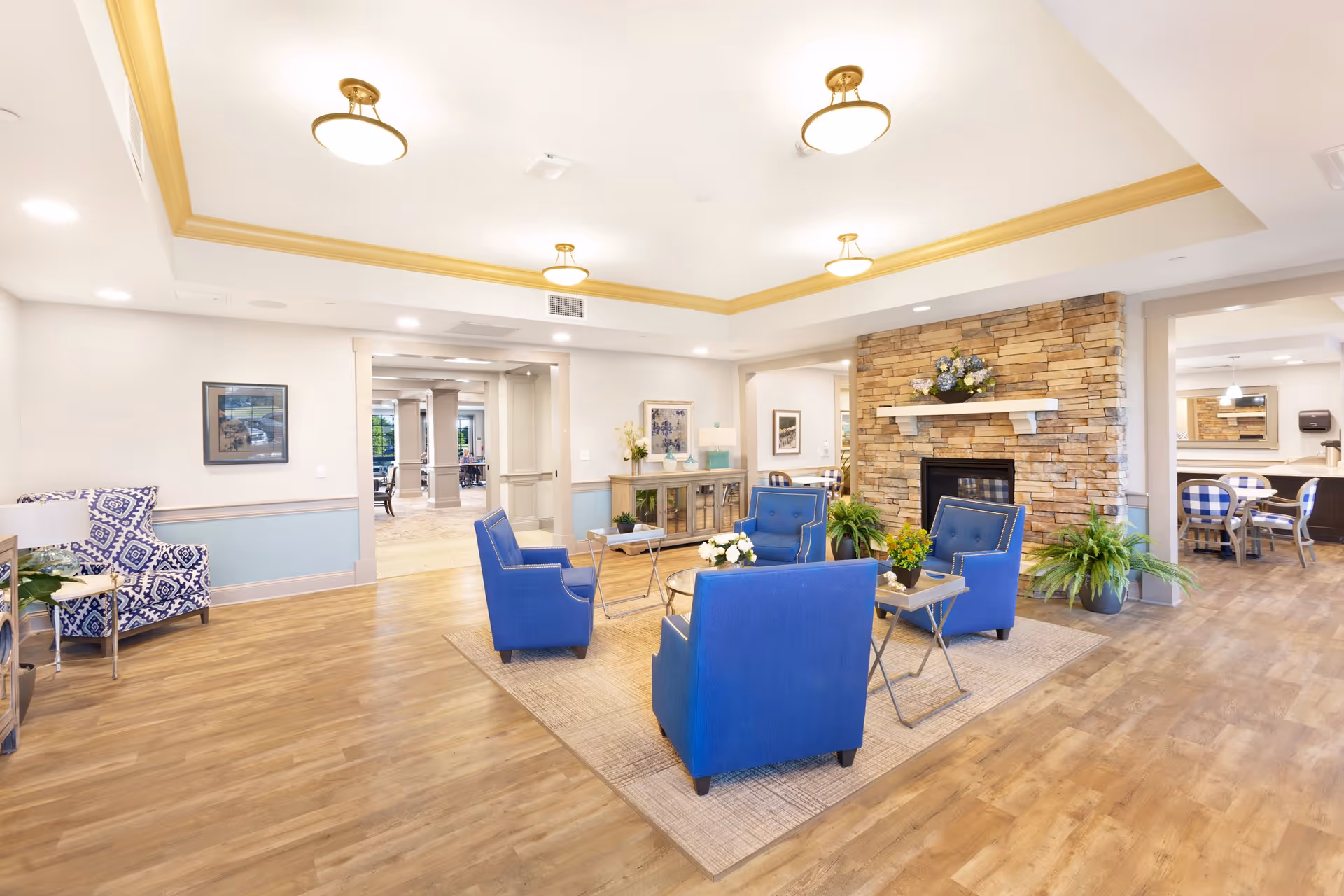A bright and spacious living room area with four blue armchairs arranged around a small coffee table on a beige rug. The room features a stone fireplace with a white mantel decorated with flowers, wood flooring, and soft lighting from ceiling fixtures. There are plants on either side of the fireplace and additional seating visible in adjoining rooms.