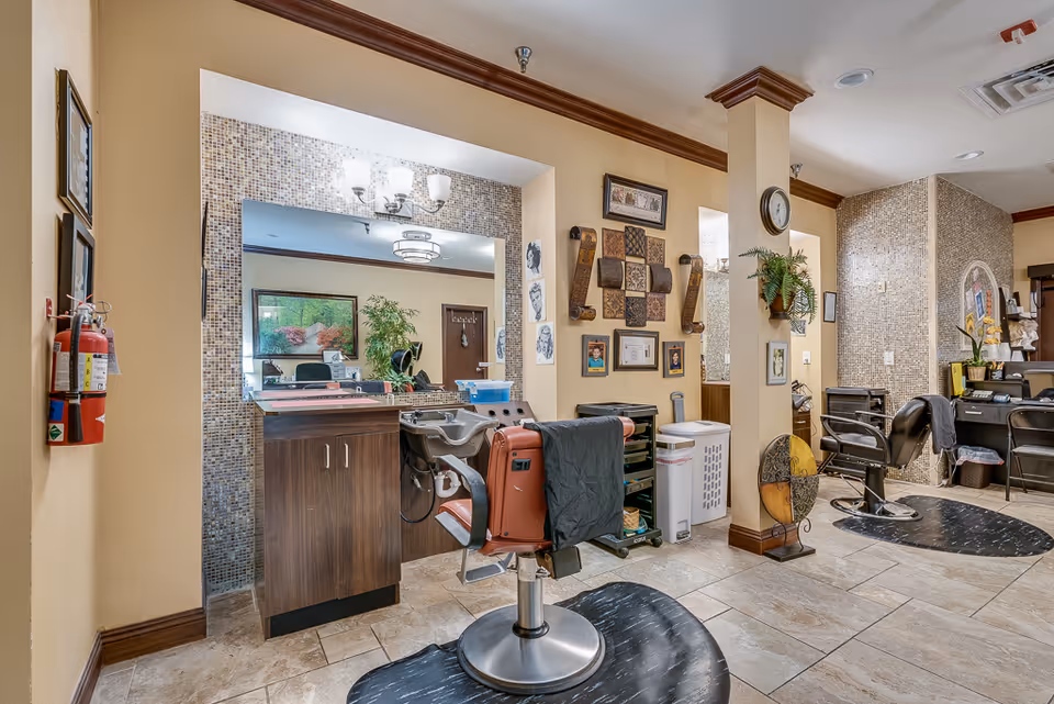 Interior view of a hair salon area within a senior living facility, featuring salon chairs, a washing station, large mirrors, decorative wall art, and plants. The space has tiled flooring, beige walls with crown molding, and bright lighting fixtures.