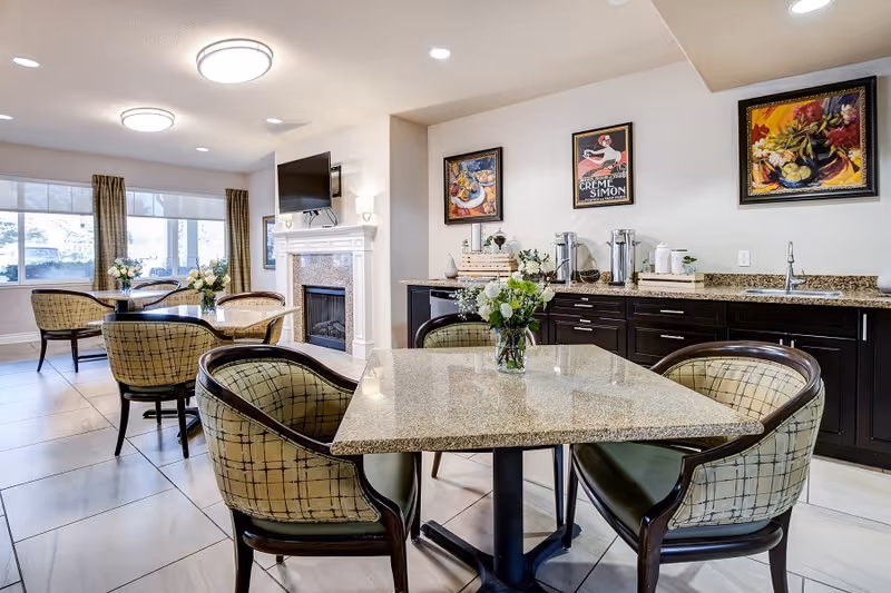A bright and clean dining area in a senior living facility with several tables and cushioned chairs. The room features a fireplace with a mounted TV above it, large windows with curtains letting in natural light, and a countertop with coffee and tea supplies. The walls are decorated with colorful framed artwork.