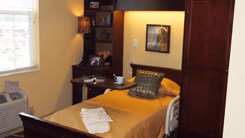 A cozy bedroom in a senior living facility with a single bed covered in a golden bedspread, a decorative pillow, and a folded white cloth at the foot. A small wooden table with a cup and an open book is placed on the bed. The room has a window with blinds, a wooden cabinet, a built-in shelf with framed photos and decorative items, a wall lamp, and a framed picture on the wall.