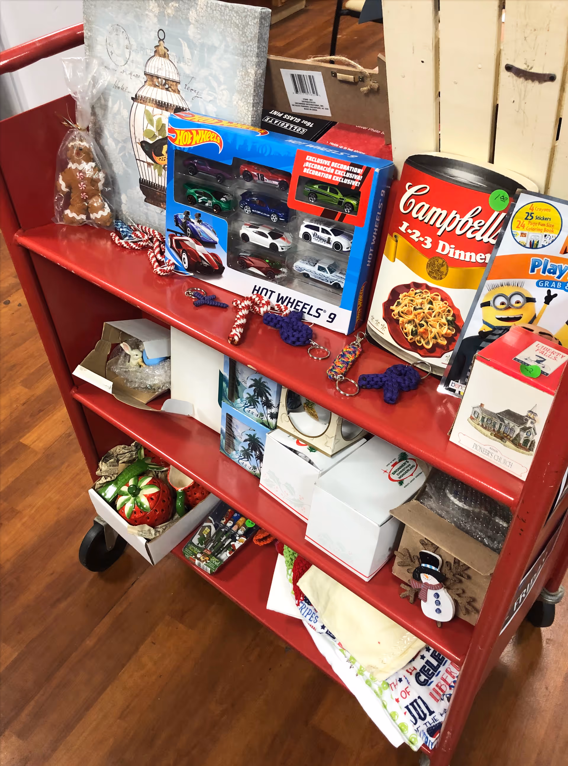 A red three-tiered cart filled with various items including a Hot Wheels toy car set, a large Campbell's soup can decoration, a gingerbread man cookie, keychains, boxed ornaments, and folded cloths on a wooden floor.