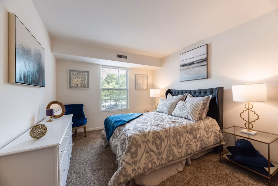 A cozy bedroom with a large bed featuring a tufted dark gray headboard and patterned bedding. There are two bedside tables with matching lamps, a white dresser with decorative items, a blue upholstered chair, and three framed artworks on the walls. A window lets in natural light, illuminating the carpeted floor.