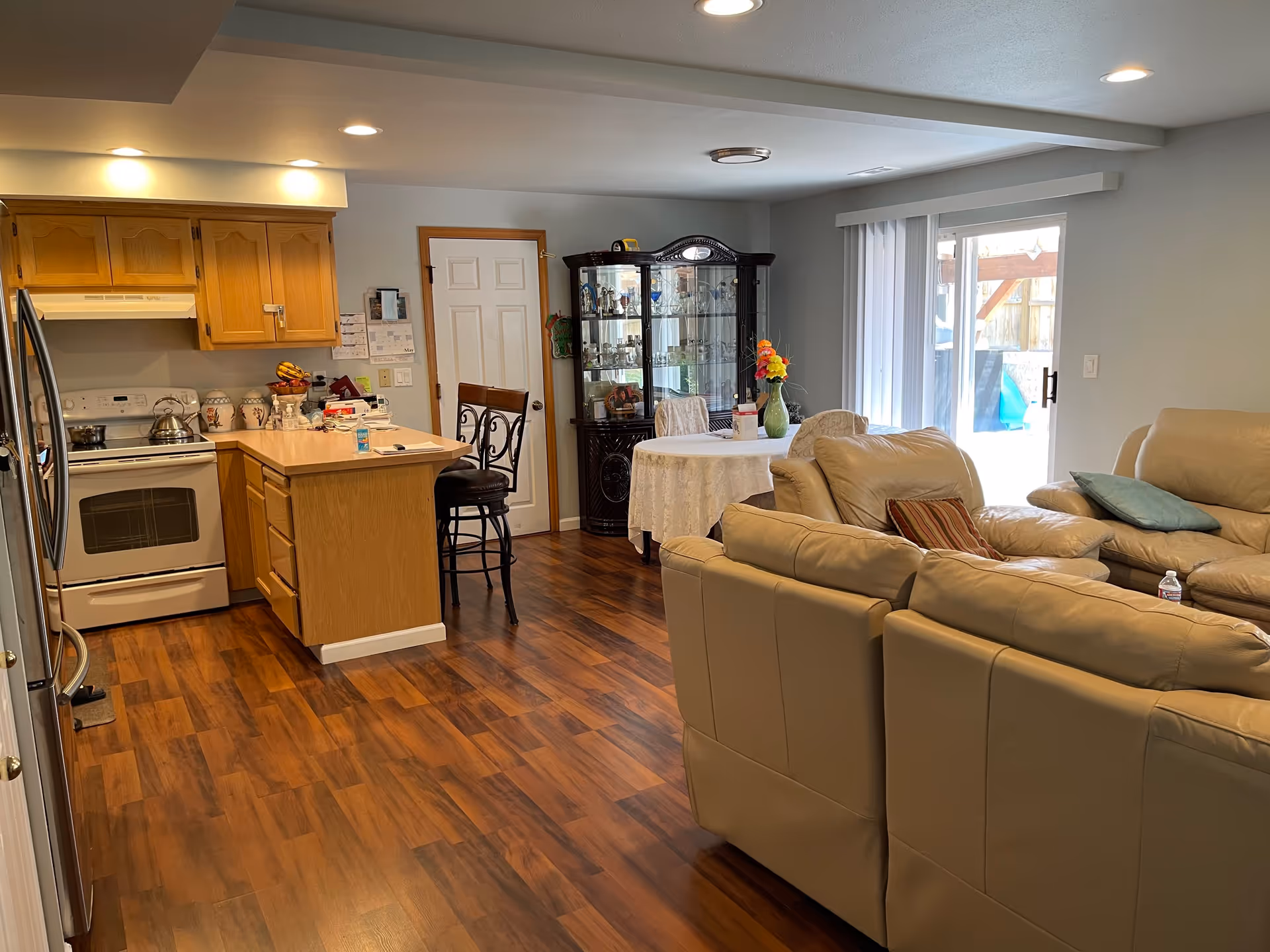 Open-plan interior showing a kitchen with an island, a round dining table with a china cabinet, and beige leather sofas in the living area.