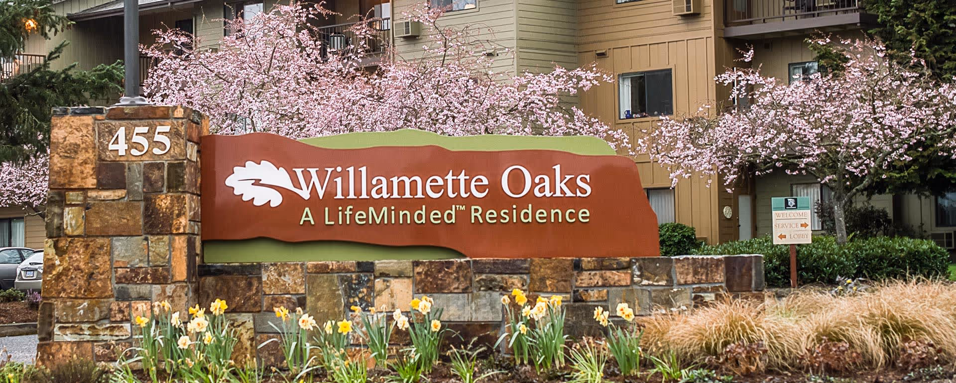 Entrance sign for Willamette Oaks, a LifeMinded Residence, surrounded by blooming pink cherry blossom trees and yellow daffodils, with a multi-story residential building in the background.