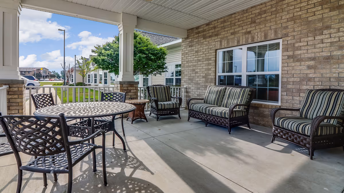 Covered outdoor patio area with metal table and chairs, cushioned wicker loveseat and armchairs, brick wall with window, and a view of green lawn and neighboring buildings under a partly cloudy sky.
