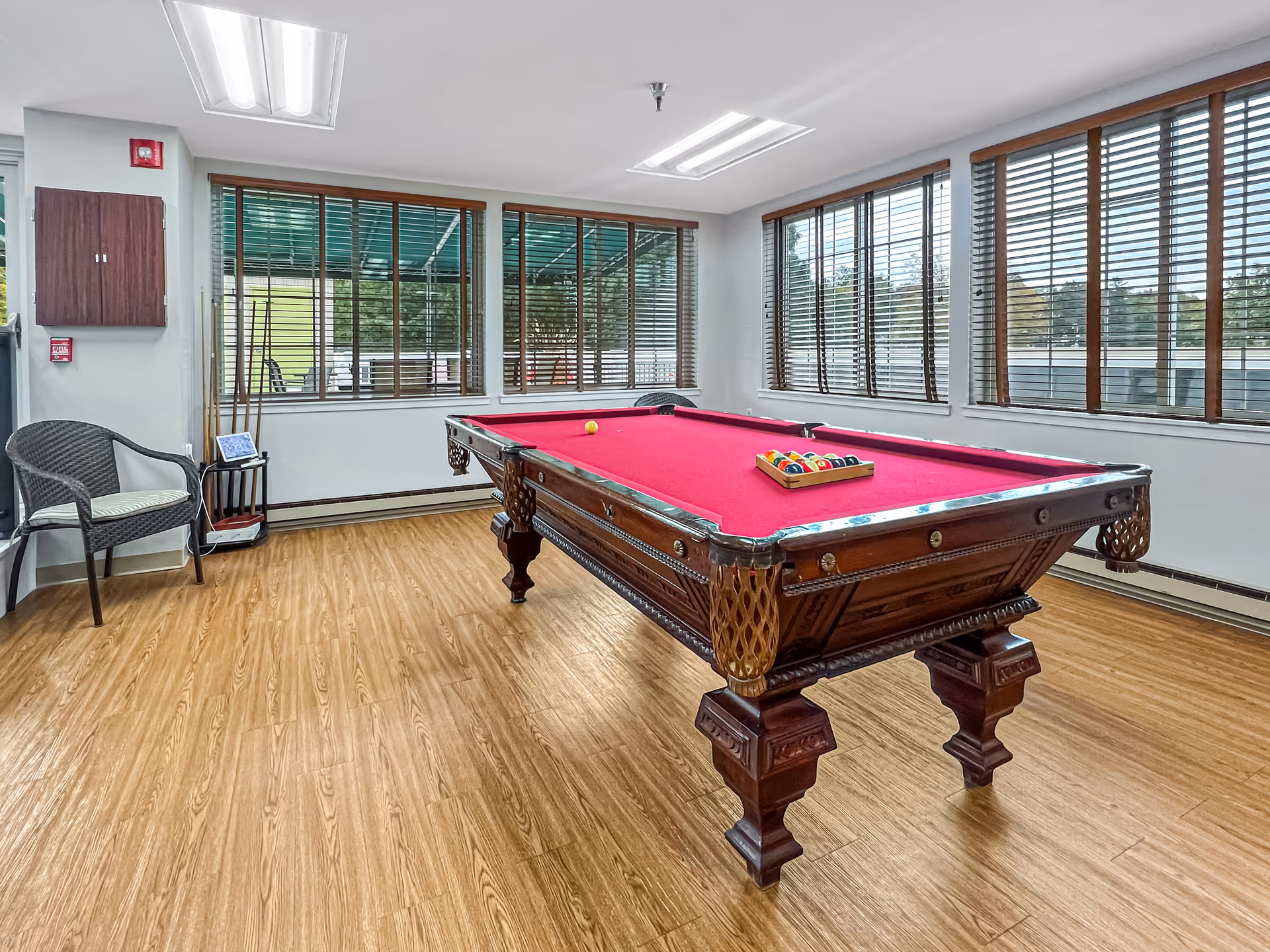 A bright room with large windows covered by wooden blinds, featuring a classic wooden pool table with a red felt surface. The pool table is set up with a rack of billiard balls and a single yellow ball on the table. There is a wicker chair with a cushion and a small rack holding pool cues in the corner. The floor is wooden, and fluorescent lights are mounted on the ceiling.