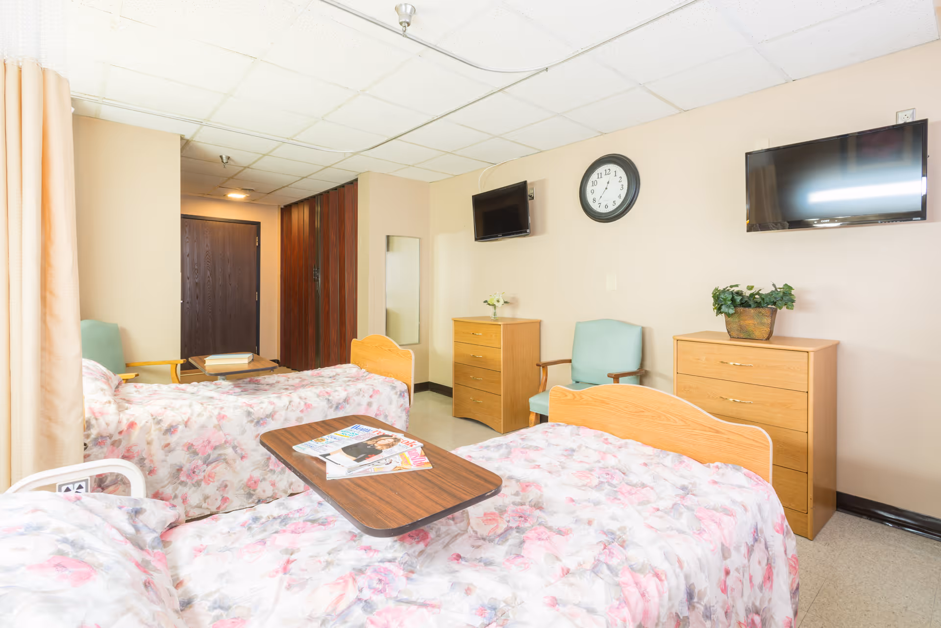 A nursing facility room with two beds covered in floral bedspreads, a wooden table with magazines between the beds, two wooden dressers, two green armchairs, a wall clock, and two wall-mounted televisions. The room has beige walls and a tiled ceiling.