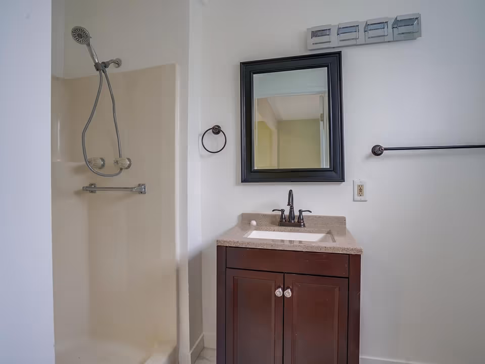 A bathroom with a shower area on the left featuring a handheld showerhead and two knobs. On the right, there is a dark wood vanity with a beige countertop, a sink, and a faucet. Above the vanity is a rectangular mirror with a dark frame. There are towel holders on the wall, one circular near the vanity and one horizontal on the right side.