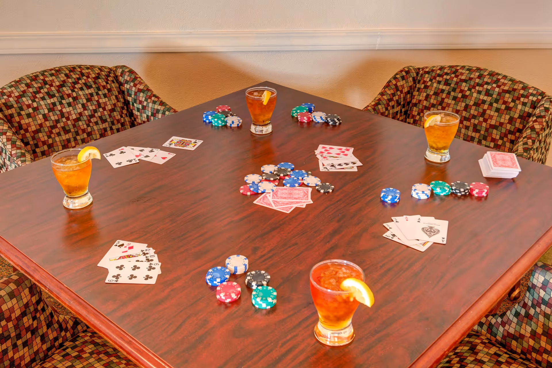 A wooden table with four patterned chairs around it, set up for a card game with playing cards, poker chips, and four glasses of iced tea garnished with lemon wedges.