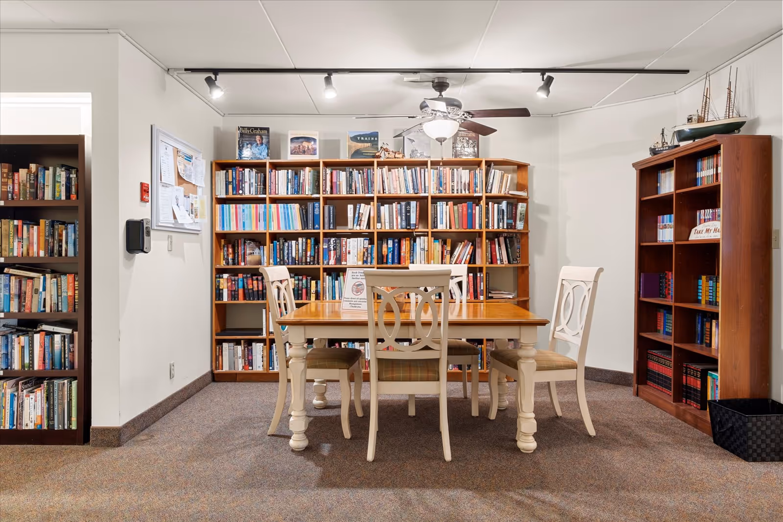 A cozy communal library room with bookshelves lining the walls and a wooden table surrounded by four chairs.