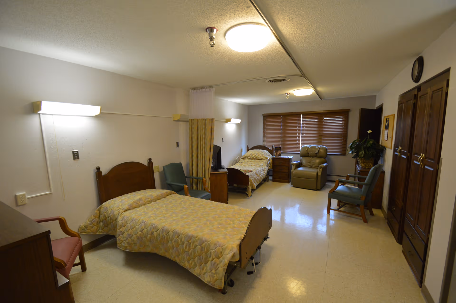 A spacious senior living shared bedroom with two hospital-style beds, chairs, a recliner, and wooden furniture under ceiling lights.